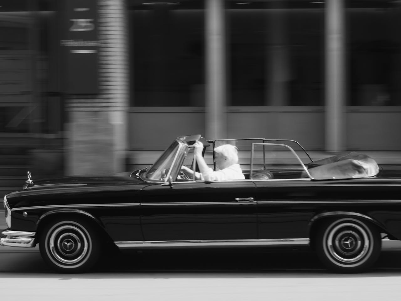 Classic black convertible with a driver cruising on an urban street in black and white.